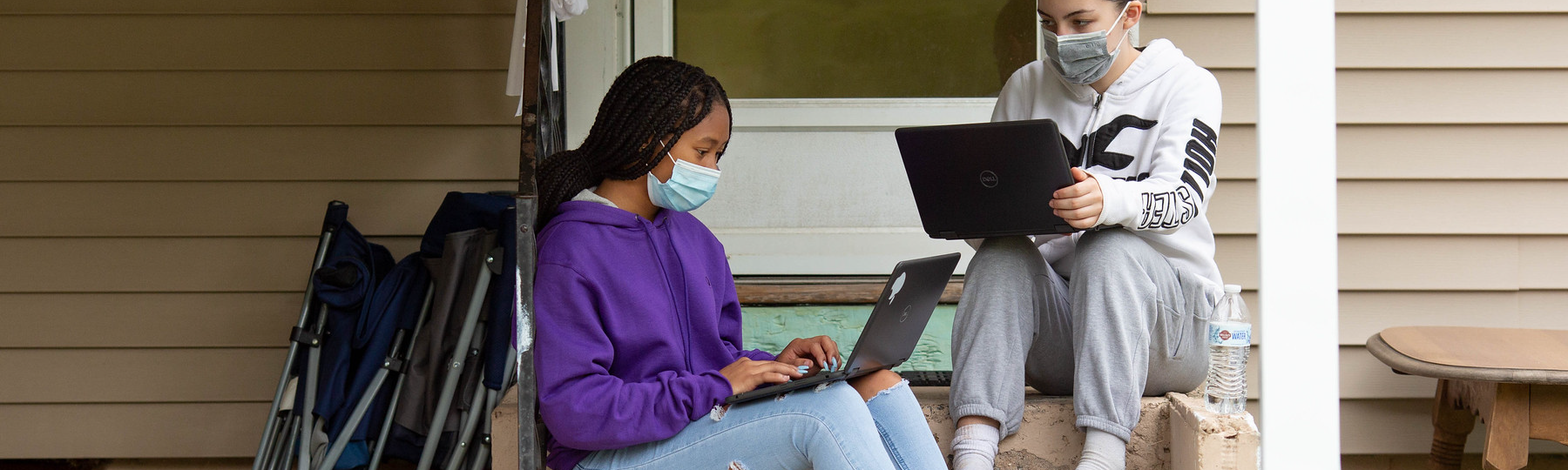Two girls who are college students sit on their front porch with face masks on. They are doing homework on their laptops.