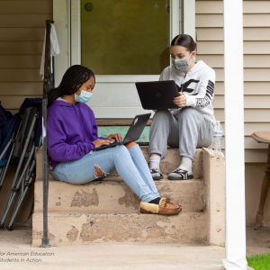 Two girls who are college students sit on their front porch with face masks on. They are doing homework on their laptops.