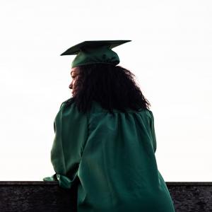 A girl in a college graduation gown stands alone, she is meant to represent Chicago Public Schools graduates who have low GPAs but deserve more equitable higher education funding to have the systematic supports she needs to graduate from college